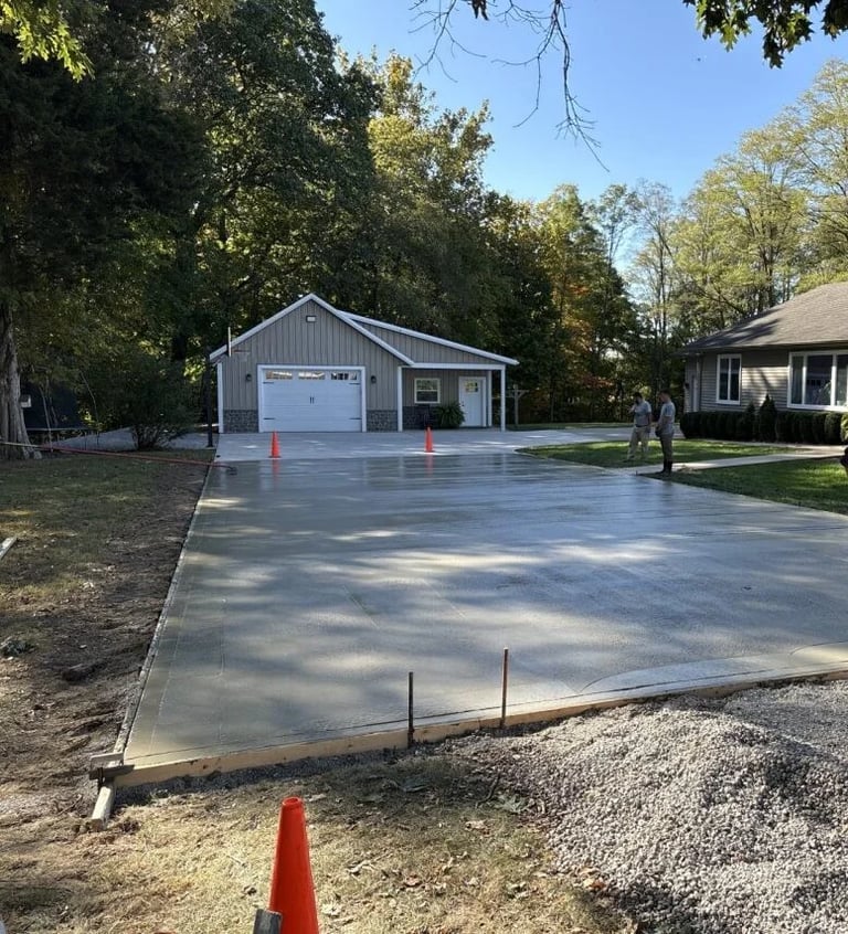 Newly poured concrete driveway with a gray garage and orange traffic cones, surrounded by trees on a residential property