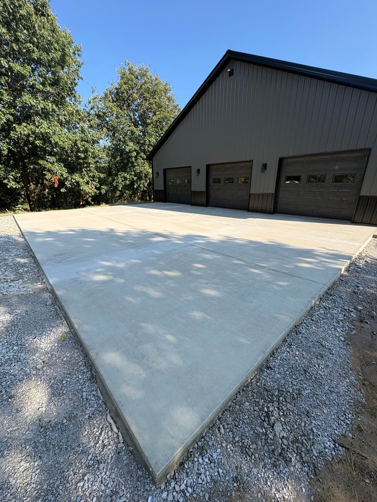 Modern black garage building with concrete driveway surrounded by gravel and green trees under clear blue sky