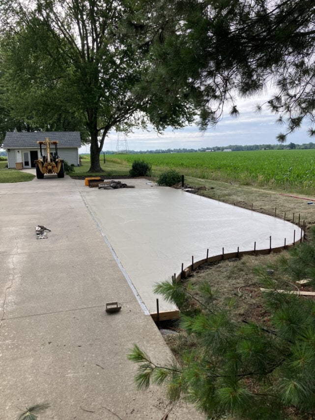 Concrete foundation pad being laid on rural farmland with construction equipment and trees nearby
