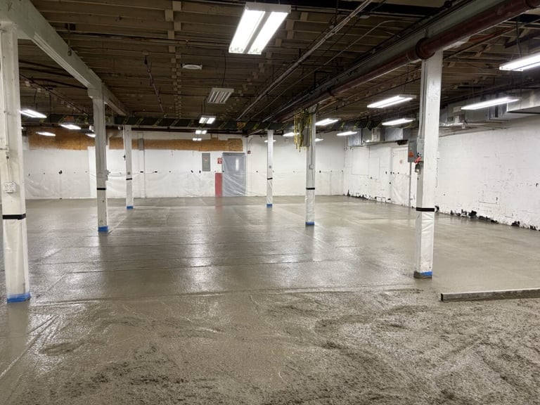 Empty industrial warehouse with concrete floor, exposed ceiling, fluorescent lights, and white support columns