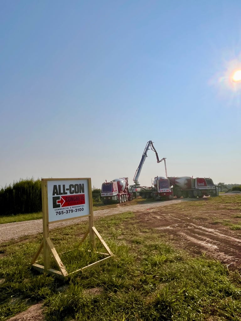 ALL-CON construction site sign in foreground with concrete pump truck and construction vehicles operating in agricultural field under clear sky