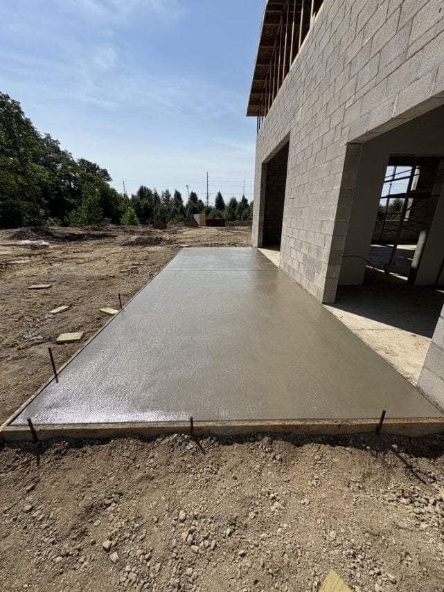 Concrete foundation pad being laid next to a gray concrete block building under construction with forest in background