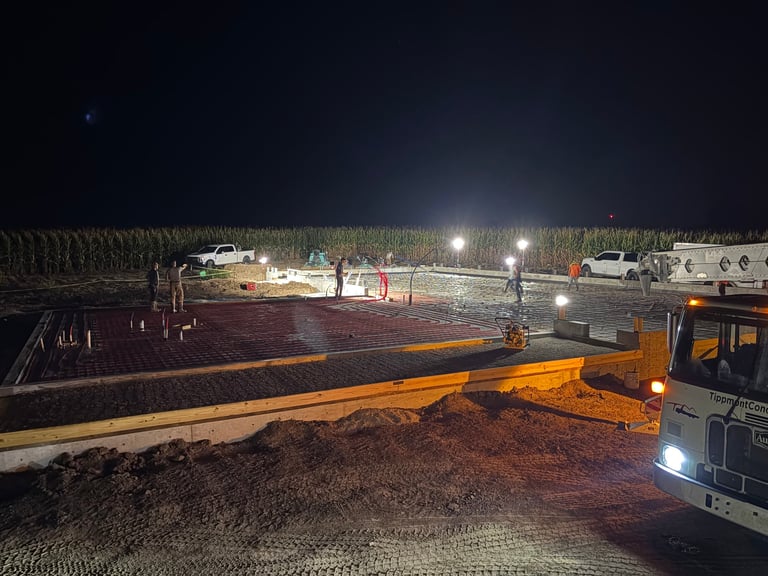 Night view of construction site with heavy machinery, yellow beam, and cornfield backdrop under floodlights