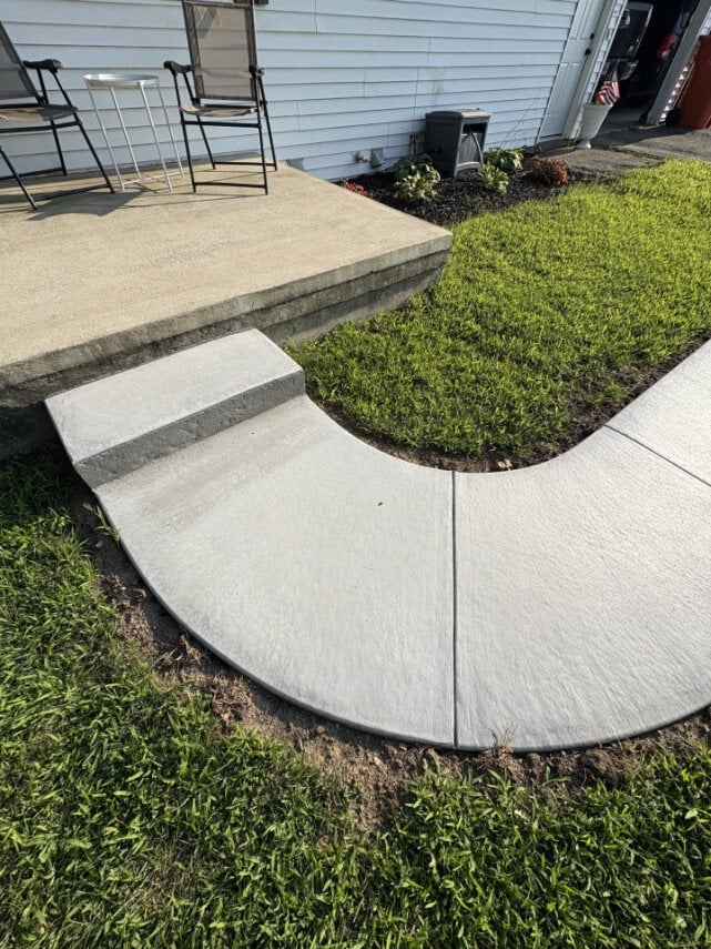 Concrete patio steps and curved walkway with green lawn, raised planter beds, and house with siding in background