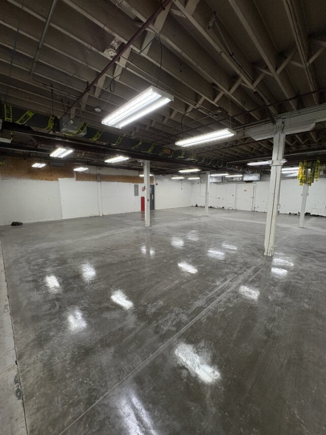 Empty industrial warehouse interior with polished concrete floor, fluorescent ceiling lights, white support columns, and metal beam structure