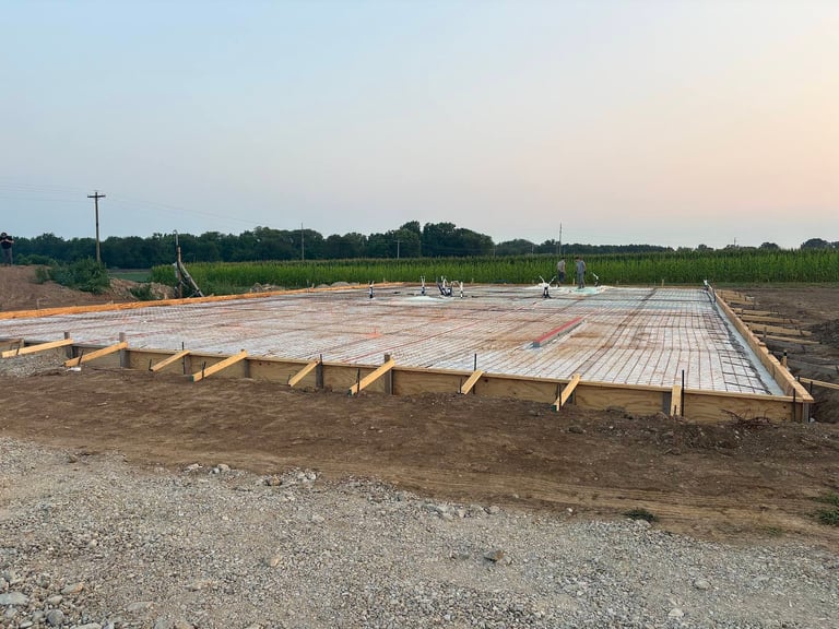 Construction site with concrete foundation pad and wooden support posts in rural field during daytime