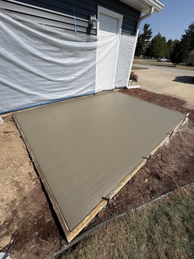 Freshly poured concrete pad next to white-draped house foundation with wood frame border and surrounding landscape