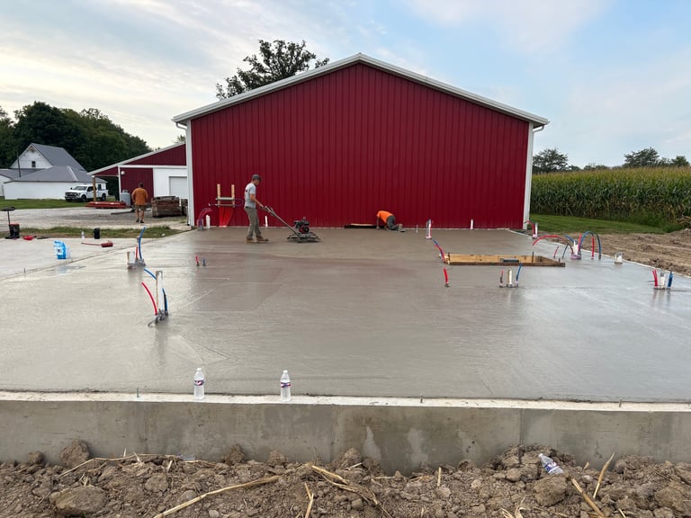 Construction site with wet concrete foundation and red barn building in background, farm setting with cornfield