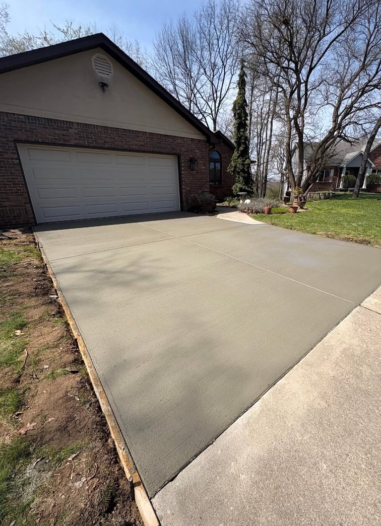 Newly poured concrete driveway in front of brick garage with bare trees and green lawn in background