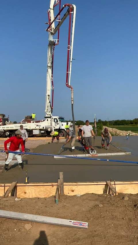 Concrete pump truck pouring gray concrete into a construction site with workers standing in the wet concrete holding finishing tools