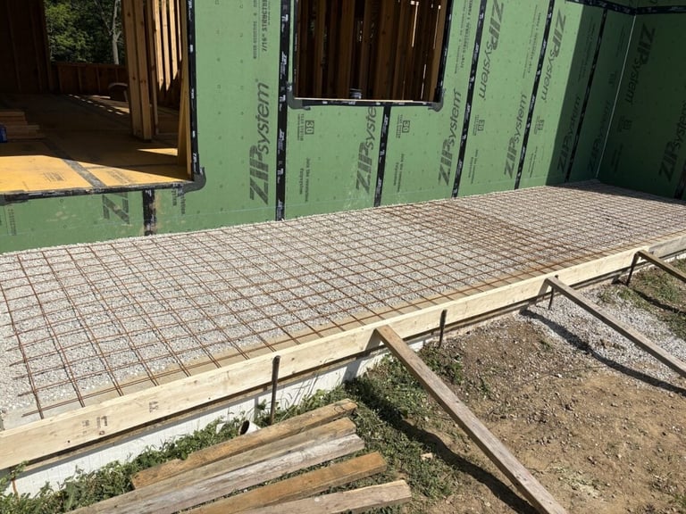Construction site showing a concrete foundation with wire mesh reinforcement and wooden framing under a green building wrap.