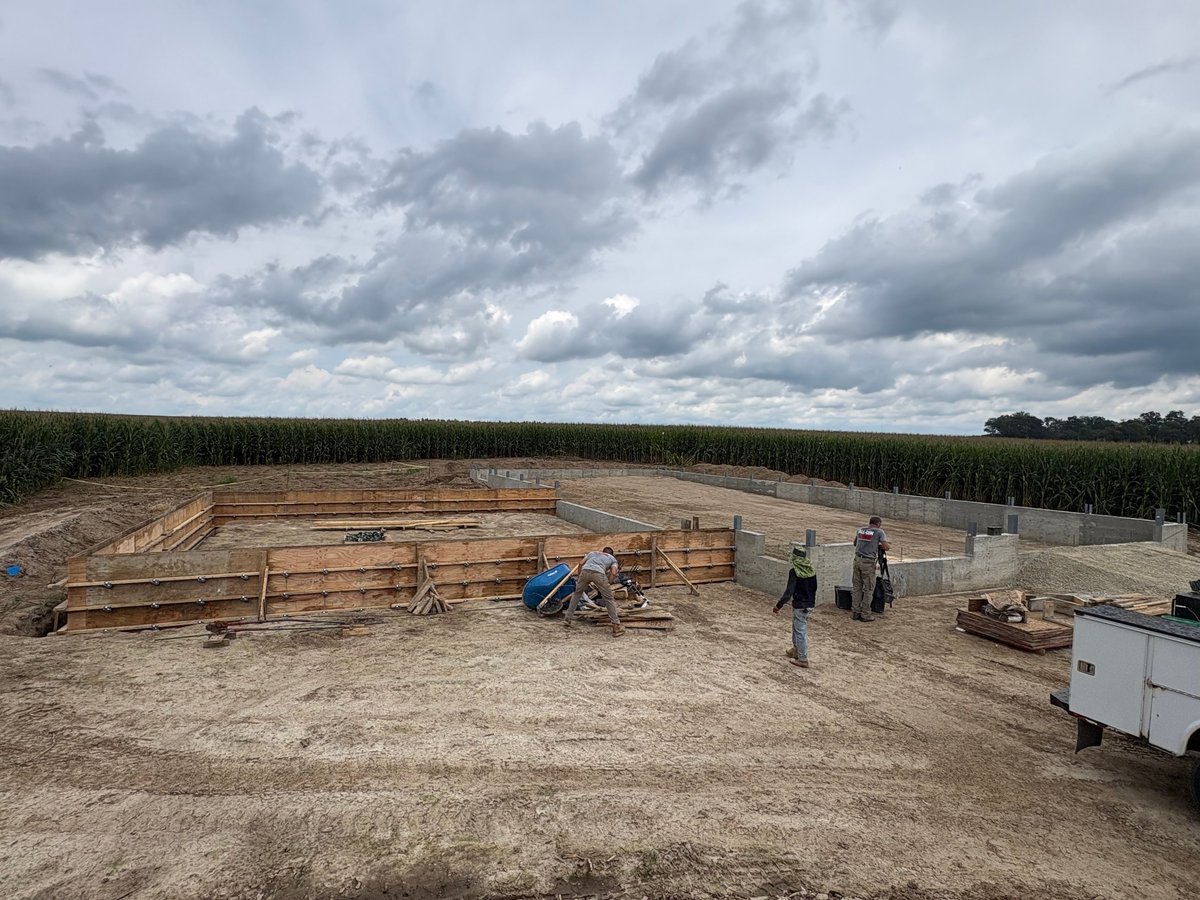 Construction site with wooden framework and workers in a cleared field surrounded by tall green crops and dramatic cloudy sky.