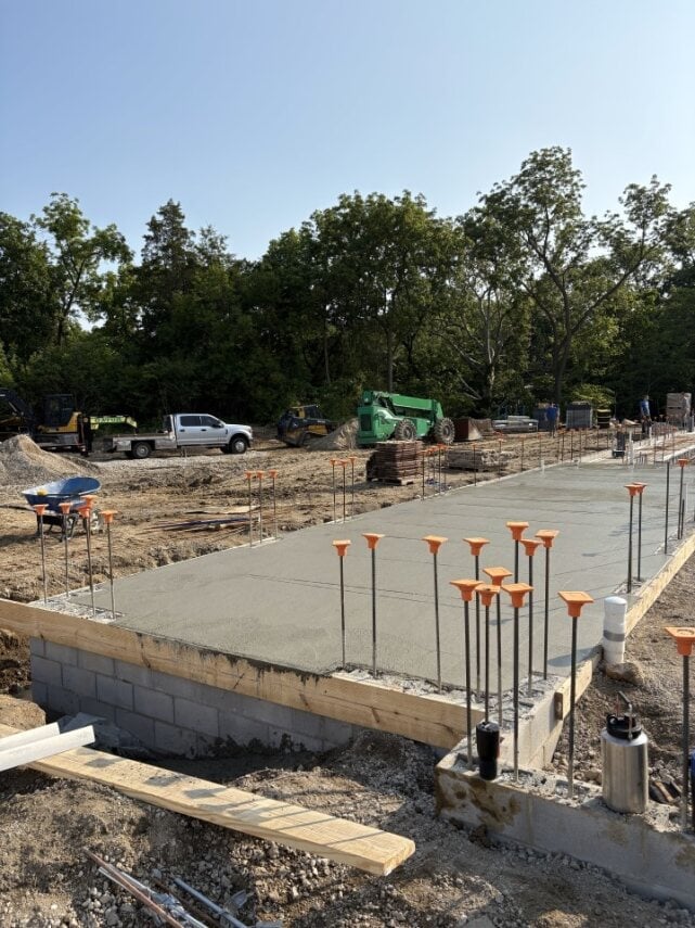 Construction site with freshly poured concrete pad, orange survey stakes, and equipment parked in background surrounded by trees