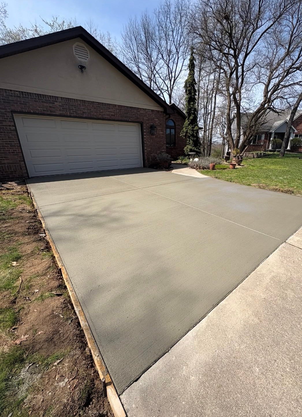 New concrete driveway in front of brick garage with bare trees and lawn visible in residential setting
