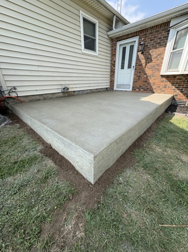 A newly poured concrete patio foundation next to a brick and siding house with a white door