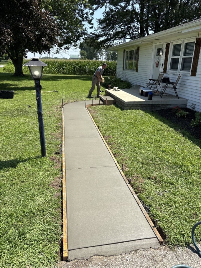 A concrete walkway leading to a white mobile home with a deck, surrounded by green lawn and trees, with a lamp post visible on the left