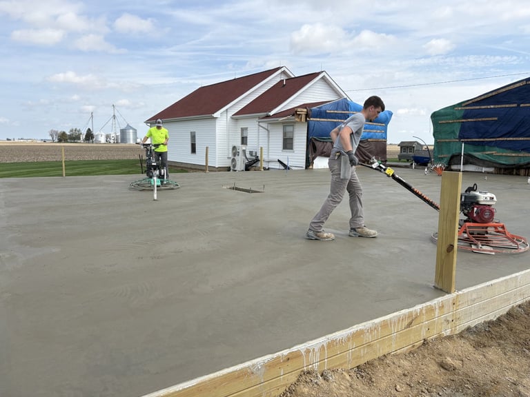 Construction workers smoothing wet concrete slab with tools, white house and equipment visible in background