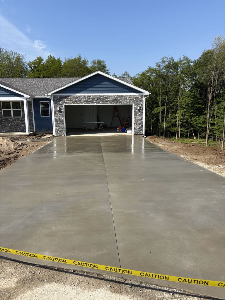 New concrete driveway with caution tape in front of a two-car garage attached to a blue and stone house with trees in background