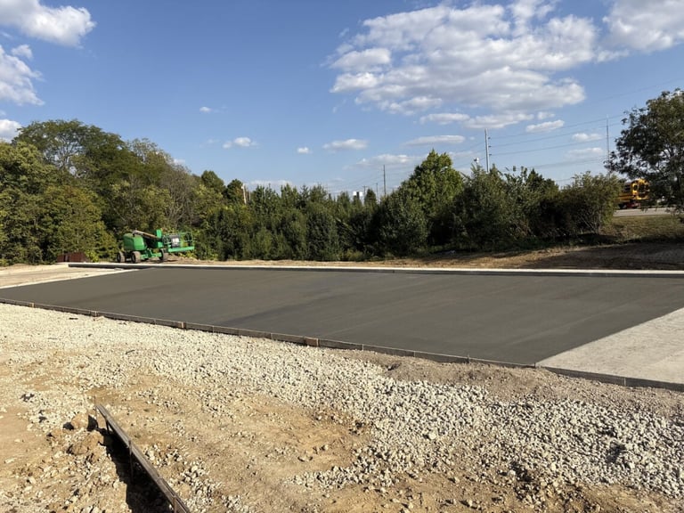 Construction site with freshly poured concrete foundation surrounded by gravel and trees under blue sky