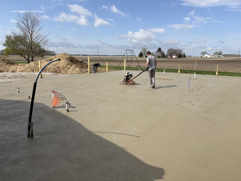 Workers finishing concrete surface at a rural construction site with farming fields visible in the background