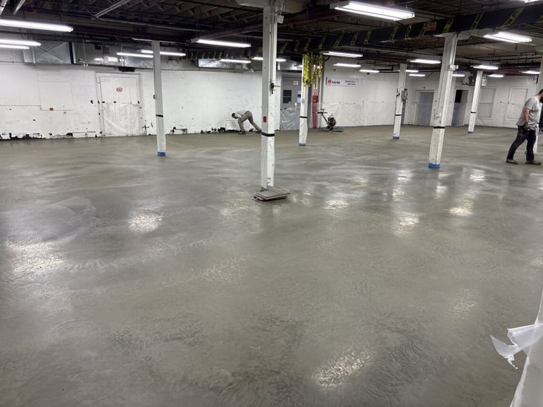 Large empty industrial warehouse with polished concrete floor, white support columns, overhead lighting, and a person walking in the distance